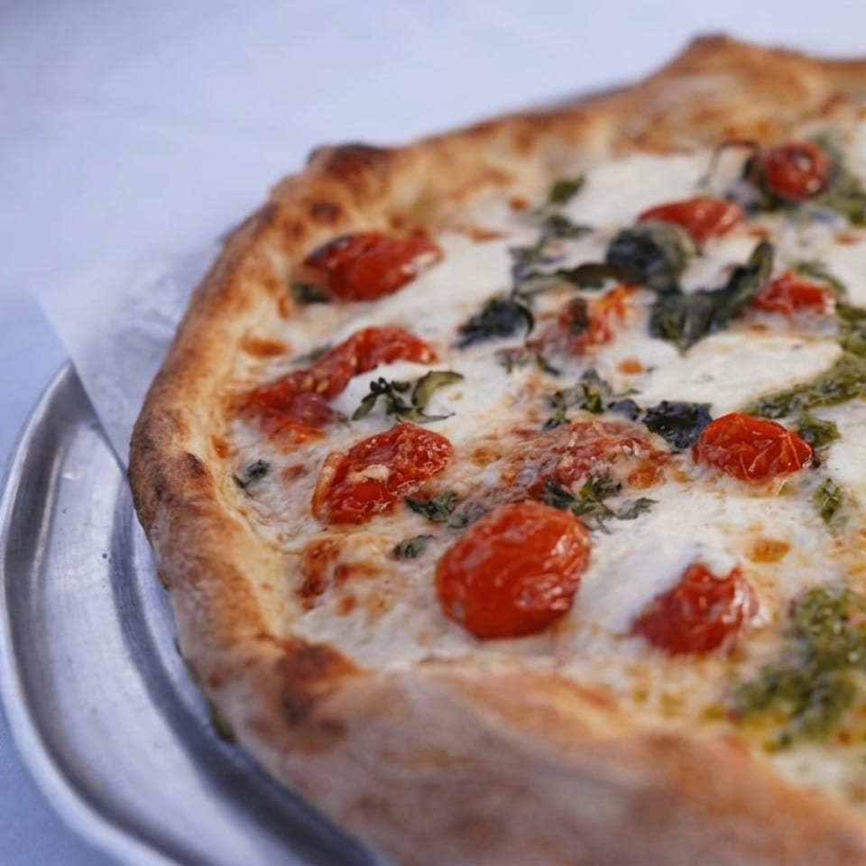 Close-up of a wood-fired pizza topped with melted mozzarella, cherry tomatoes, basil, and olive oil on a metal plate.