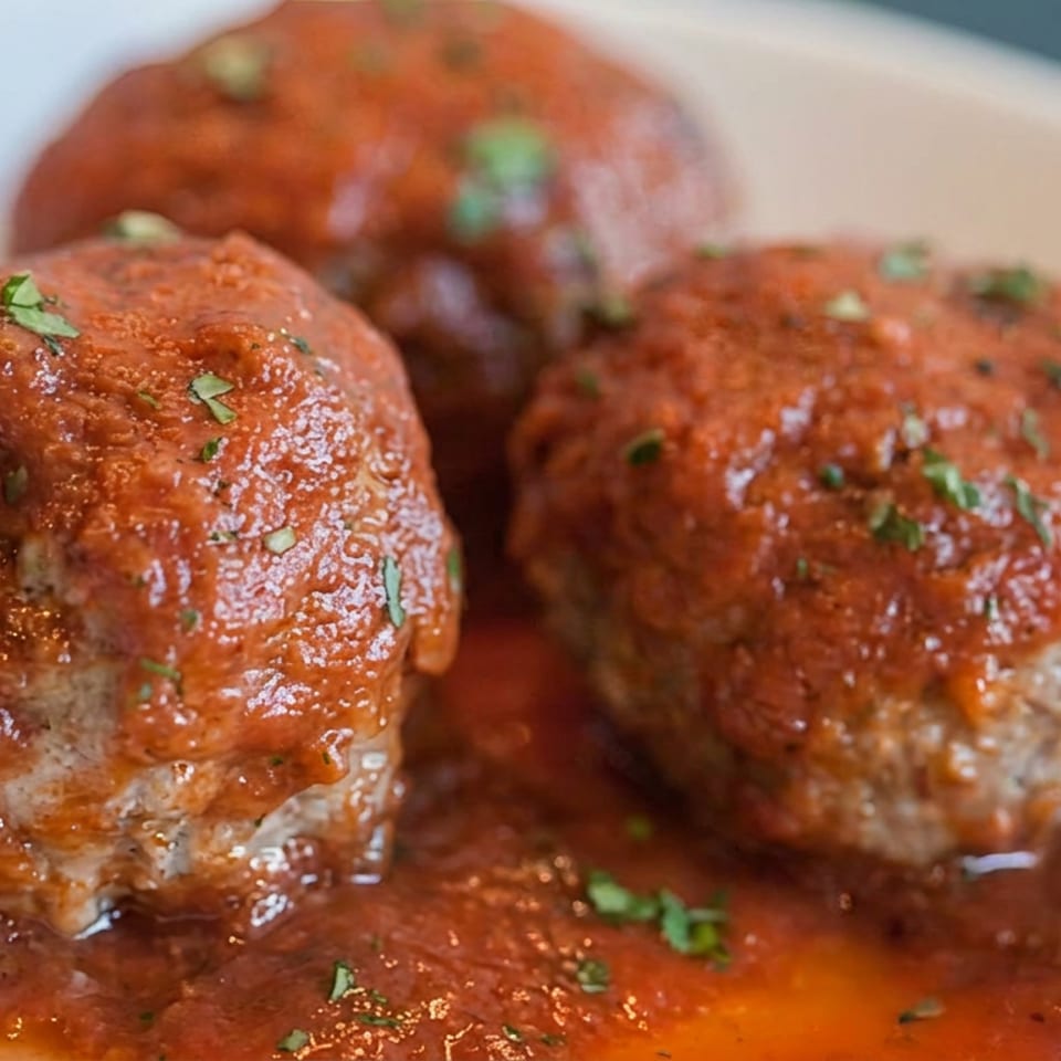 Three meatballs covered in tomato sauce and sprinkled with fresh herbs on a white plate.