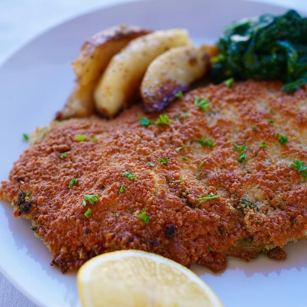 Crispy breaded chicken cutlet with roasted potatoes, spinach, and a lemon wedge on a white plate.